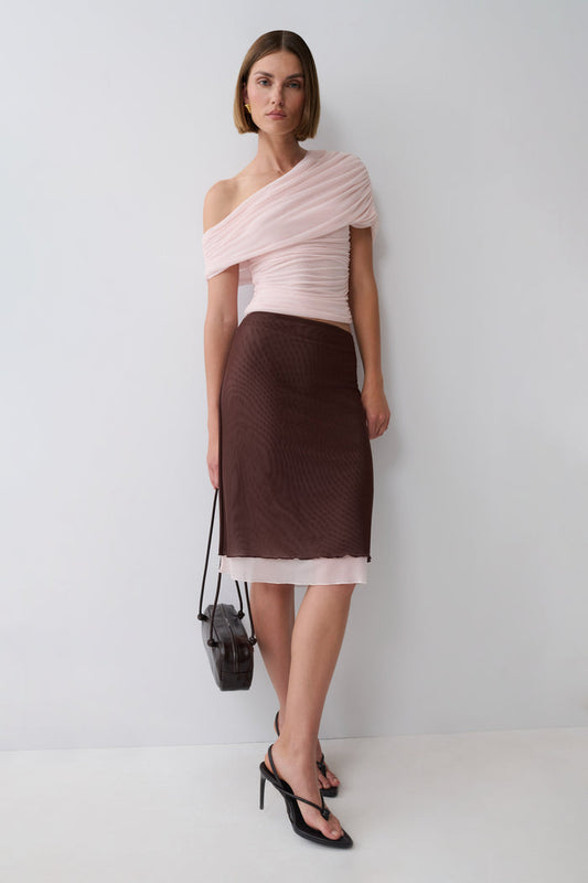A model stands against a light gray studio wall wearing a pale pink one-shoulder top and a dark brown textured pencil skirt with a sheer pink underlayer. She is holding a small black handbag and wearing black strappy heels.