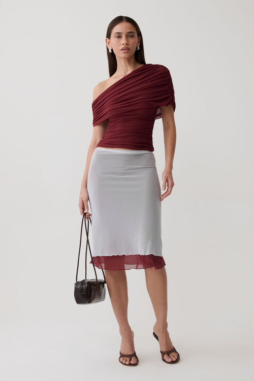 A model stands against a light gray studio background wearing a draped off-the-shoulder burgundy top and a fitted white pencil skirt with a sheer burgundy hem, holding a small black handbag.