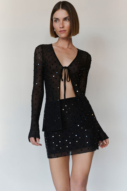 A model wearing a black sheer long-sleeve top and matching mini skirt set, both embellished with small iridescent sequins, against a plain light gray background.
