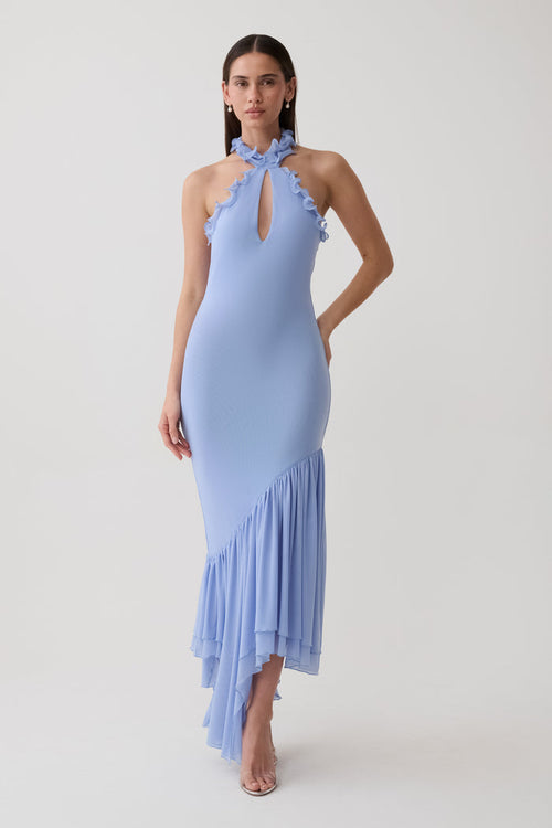 A woman modeling a light blue halter-neck maxi dress with ruffle trim and an asymmetrical ruffled hem, photographed against a plain light gray studio background.