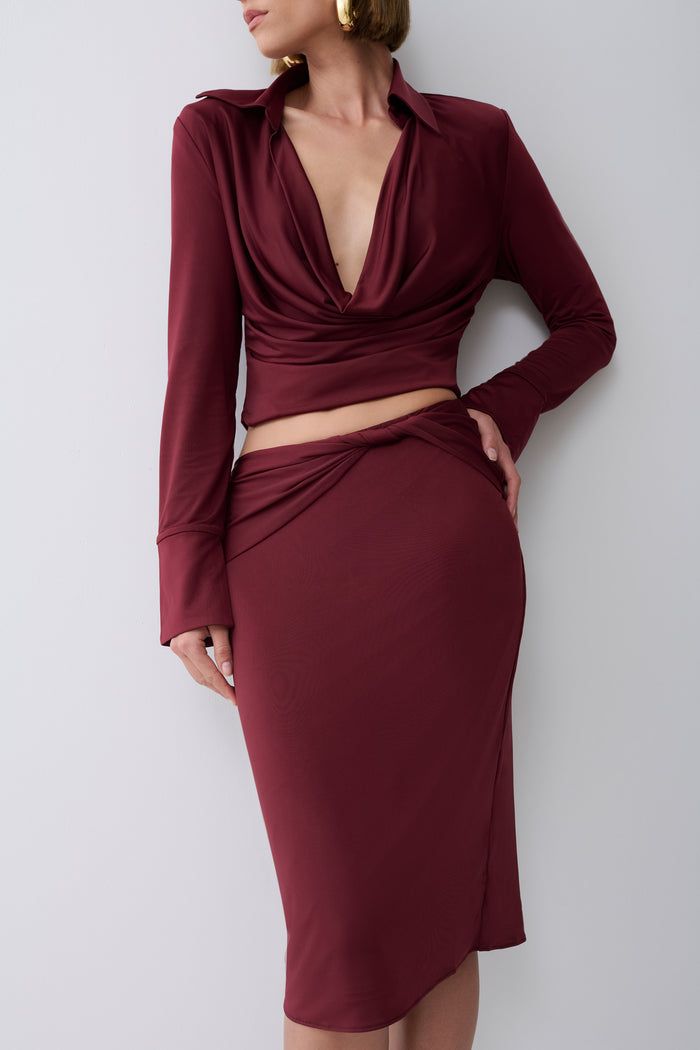 A woman modeling a matching burgundy two-piece set consisting of a long-sleeve cowl-neck crop top and a ruched midi skirt, photographed against a plain light gray studio background.