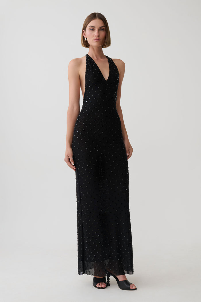 A woman modeling a sleeveless black halter-neck maxi dress with a subtle dotted pattern, photographed against a plain light gray studio background.